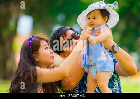 young happy and loving Asian Japanese parents couple enjoying together with sweet daughter baby girl sitting on grass at green city park in love and b Stock Photo