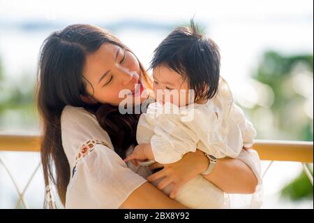 young happy and beautiful Asian Korean woman playing and holding sweet adorable baby girl sitting at holidays resort enjoying summer vacation trip in Stock Photo
