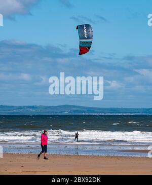 Firth of Forth, Scotland, United Kingdom, 6th June 2020. UK Weather: a kite surfer returns to the sea after lockdown easing allows some outdoor sports. After a dull start on a stormy day the sun came out to create just the right conditions at Broadsands Bay as a woman walks along the beach