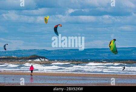 Firth of Forth, Scotland, United Kingdom, 6th June 2020. UK Weather: a group of kite surfers return to the sea after lockdown easing allows some outdoor sports. After a dull start on a stormy day the sun came out to create just the right conditions at Broadsands Bay as a woman walks along the beach