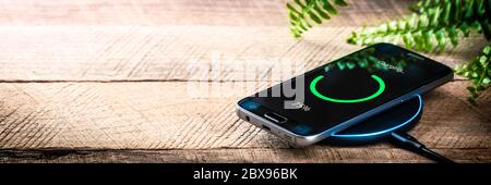 Smartphone On Wireless Charging Station On Wooden Desk With Green Plant Stock Photo