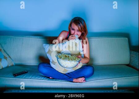 young beautiful and scared woman watching horror or suspense movie eating popcorn sitting at home sofa couch late night in panic and fear facial expre Stock Photo