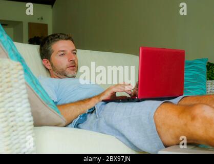 lifestyle indoors portrait of young attractive and happy man lying at home sofa couch relaxed using internet on laptop computer watching online movie Stock Photo