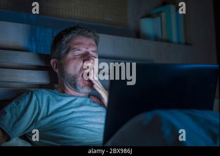 attractive tired and stressed workaholic man working late night exhausted on bed busy with laptop computer yawning feeling sleepy and overworked in bu Stock Photo