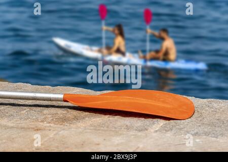 Close-up of a paddle for kayak on the quay with two people out of focus, paddling in the sea on background Stock Photo