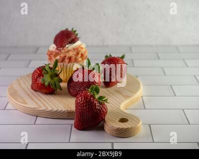 Strawberry shortcake cupcake in the background with 4 strawberries in front of it, arranged on a round wooden cutting board with a handle on a white s Stock Photo