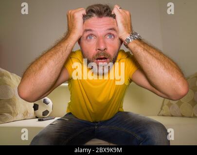 close up expressive portrait of young excited and nervous football supporter man watching soccer game on television at living room sofa feeling crazy Stock Photo