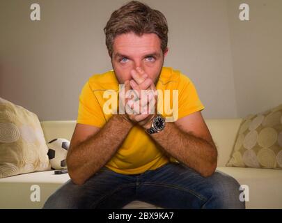 close up expressive portrait of young excited and nervous football supporter man watching soccer game on television at living room sofa feeling crazy Stock Photo