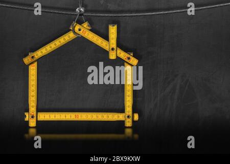 Yellow wooden folding ruler in the shape of a house hanging on a steel cable, on a blackboard with copy space
