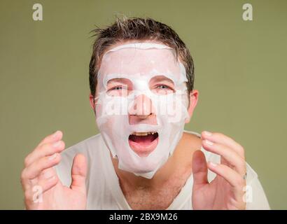 natural portrait of young happy and funny man applying beauty facial mask looking in the mirror laughing cheerful finding himself weird and scary isol Stock Photo
