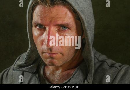 close up sweaty face portrait of young attractive and fierce looking man wearing hoodie posing in aggressive and defiant attitude isolated on dark bac Stock Photo