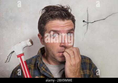 portrait of man holding hammer driving a nail for hanging a frame but making funny faces for the  mess cracking the wall as a disaster DIY guy and mes Stock Photo