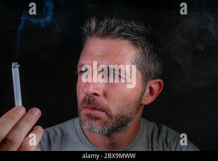 close up portrait of young handsome and thoughtful tobacco addict man holding cigarette thinking time to quit smoking isolated on black background in Stock Photo