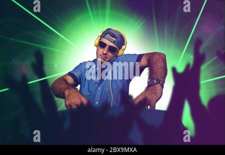 young happy and cool DJ playing music at party event in night club mixing techno songs on laser and flash lights background cheered with hands by club Stock Photo