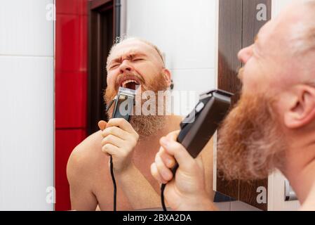 Handsome cheerful bearded man singing into hair trimmer instead of microphone in front of mirror in bathroom at home. Positive happy man joking Stock Photo