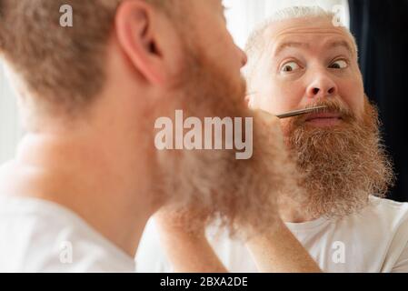 Attractive middle-aged man cutting his moustache and beard by himself in front of mirror at home. Beard care during quarantine