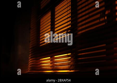 Dark, moody atmosphere set by yellow light softly piercing through the blinds. The shutters create a lovely contrast between light and shadow. Stock Photo