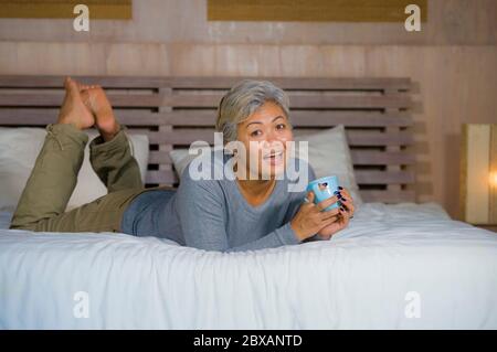 home portrait of attractive and successful mature woman with grey hair sitting on bed drinking coffee relaxed smiling happy and charming as middle age Stock Photo