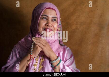 isolated studio portrait of happy and positive senior muslim woman in her 50s wearing traditional Islam hijab head scarf praying holding prayer beads Stock Photo