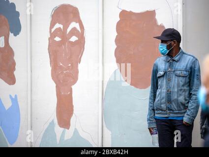 Man, wearing a face mask, baseball cap and denim jacket, stands in front of a painted wall mural, at the BLM UK protest, Whitehall, London UK Stock Photo