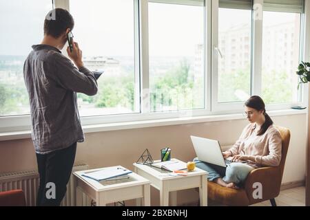 Caucasian businessman discussing on phone while his girlfriend is working at the laptop and sitting in armchair Stock Photo