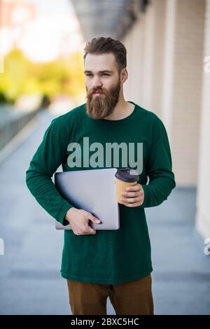 Young bearded hipster man holding laptop walking on the sunny street. Stock Photo