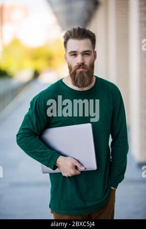 Young bearded hipster man holding laptop walking on the sunny street. Stock Photo