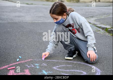 A girl with a face mask is drawing a heart on the pavement with a chalk. She is wearing a face mask due to the coronavirus pandemic. Stock Photo