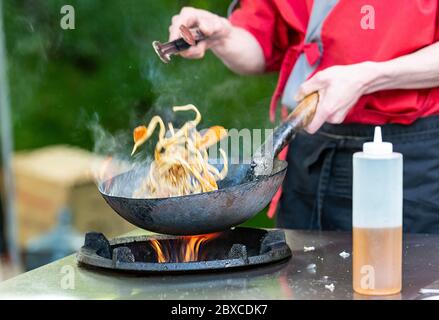 Vegetables fry on a large frying pan on fire. Cooking at the festival. Girl  chef pouring vegetables into disposable utensils. Stock Photo