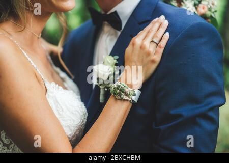 Marriage couple embracing. Bride in white wedding dress holding hand on groom's shoulder. Flower bridal accessories: bracelet and boutonniere. Stock Photo