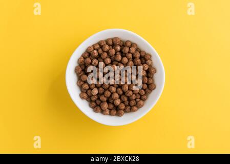 White bowl full of chocolate corn cereal balls on yellow background, top-down view. Modern vivid breakfast image. Stock Photo