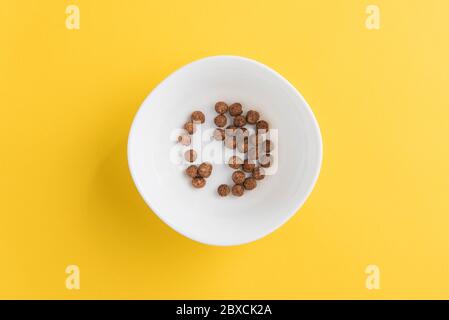White bowl with few chocolate corn cereal balls on yellow background, top-down view. Modern fresh breakfast image. Stock Photo