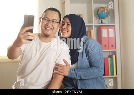 Portait of happy Asian muslim couple smiling taking selfie picture on smart phone, husband and wife hugging full of love, family concept Stock Photo
