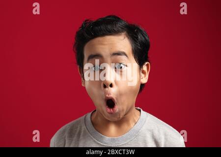 Portrait of funny young Asian boy looking at camera with open mouth and big eyes, shocked surprised expression against red background Stock Photo