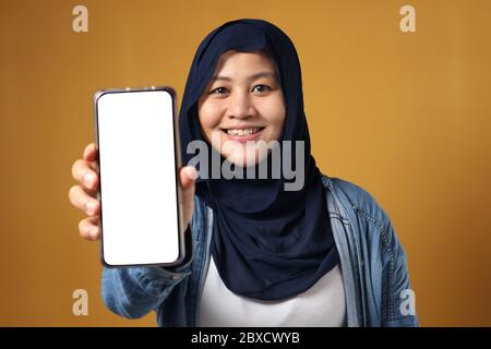 Portrait of Asian muslim woman smiling  at camera and showing her smart phone, phone mock up, against yellow background Stock Photo
