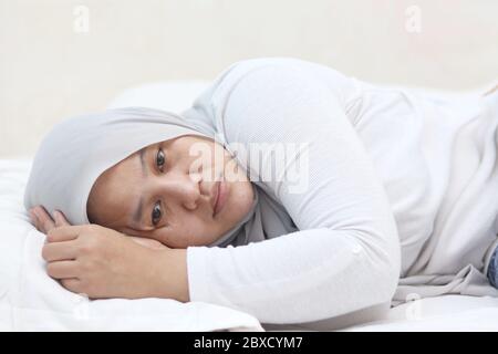 Sad unhappy Asian muslim woman lying on bed with blank stare, hard to sleep thinking of bad situation, emotional expression Stock Photo