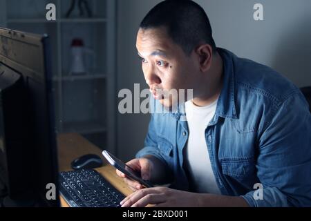 Young Asian man using smart phone and computer until late night, he is working or studying hard to reach his ambition. Student or businessman works ha Stock Photo