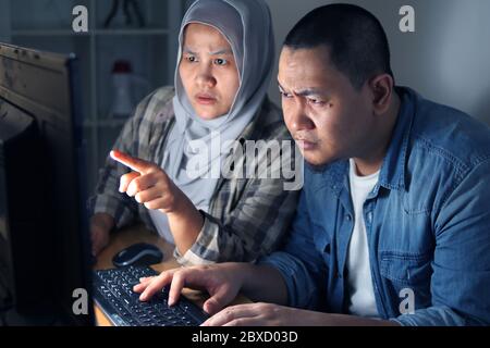 Serious muslim Asian man and woman working at night on computer. Focused professional male and female business people using laptop in dark office, web Stock Photo