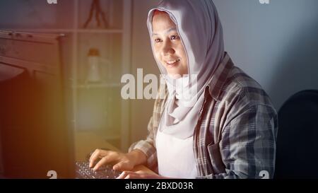 Young asian muslim female entrepreneur freelancer working at home in the middle of the night, smiling happy hard worker looking for success Stock Photo
