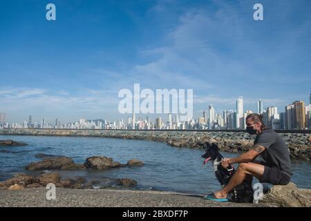 Balneario  Camboriu, Santa Catarina, Brazil - May 30, 2020: Brazilian man sitting on the seashore with pet dog wearing protective mask Stock Photo