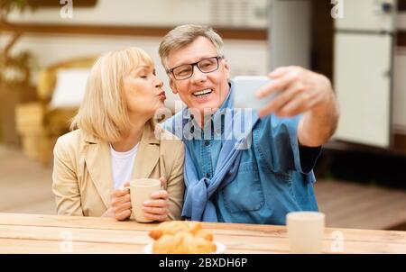 Mature woman kissing her husband while he taking selfie near motorhome outdoor Stock Photo