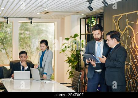 Serious young bearded businessman showing financial chart on tablet computer to colleague Stock Photo