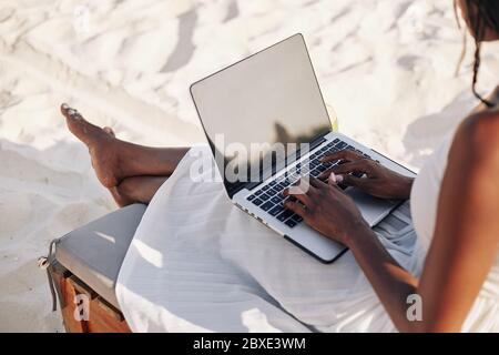 Young woman with laptop sitting on chaise-lounge on beach and working remotely Stock Photo