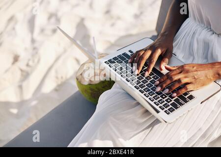 Close-up image of woman sitting on sandy beach and working on laptop, coding or answering e-mails Stock Photo