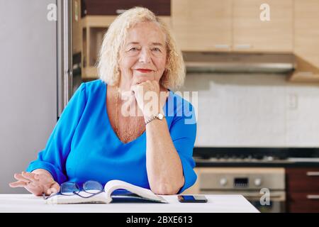 Portrait of happy senior woman sitting at kitchen table with opened book in front of her Stock Photo