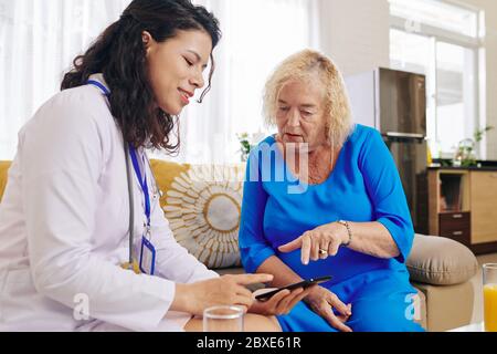 Smiling physician visiting senior patient at home and explaining her how to using healthcare application to connect with doctor Stock Photo