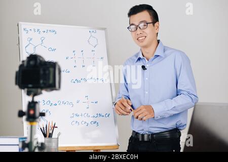 Smiling Vietnamese school teacher conducting online class and explaining formulas on whiteboard Stock Photo