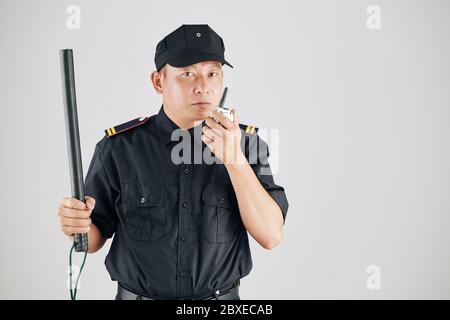 Studio photo of serious police officer raising hand with truncheon and talking to chief of police via walkie-talkie Stock Photo