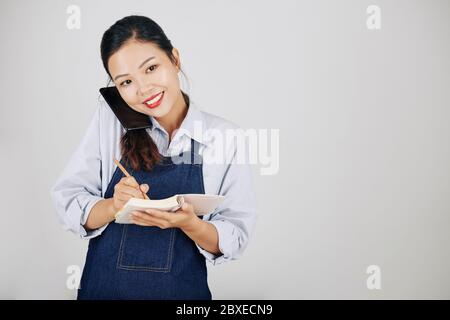 Smiling pretty Vietnamese waitress in denim apron talking on phone with customer and writing down order Stock Photo