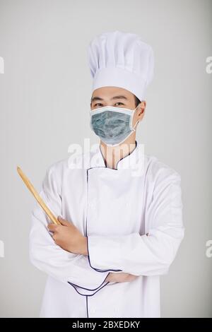 Portrait of positive restaurant chef in medical mask holding wooden spoon Stock Photo